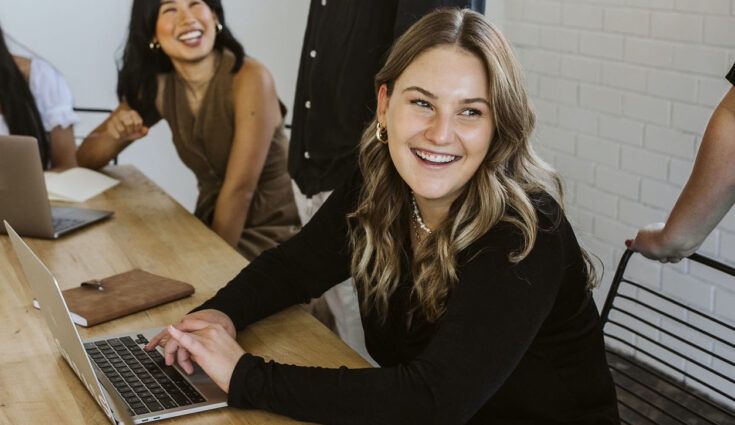 women sitting at desk, typing on laptop, smiling away from camera